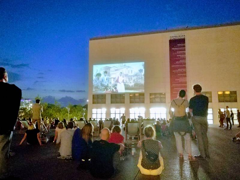 Film wird auf eine Fassade projiziert, Menschen sitzen und stehen davor und schauen zu.